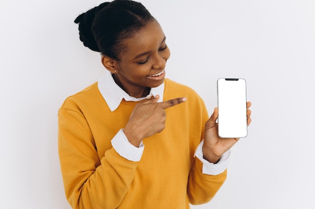 Teléfono móvil de moda. Mujer afroamericana sonriente sosteniendo el último teléfono inteligente delgado con pantalla en blanco y apuntándolo.