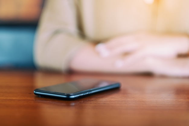 Un teléfono móvil negro sobre la mesa con una mujer sentada en segundo plano.