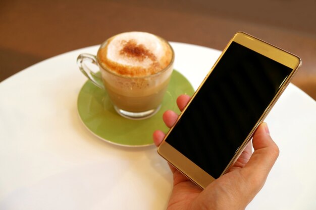 Teléfono móvil de pantalla en blanco en la mano con café capuchino en mesa redonda blanca
