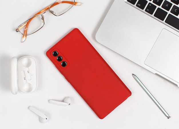 Teléfono móvil rojo, auriculares y gafas cerca de la computadora portátil sobre fondo blanco.