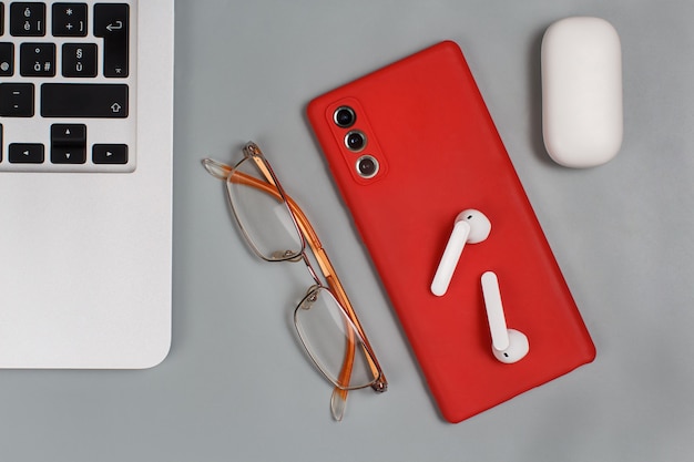 Teléfono móvil rojo, auriculares y gafas cerca de la computadora portátil sobre fondo gris