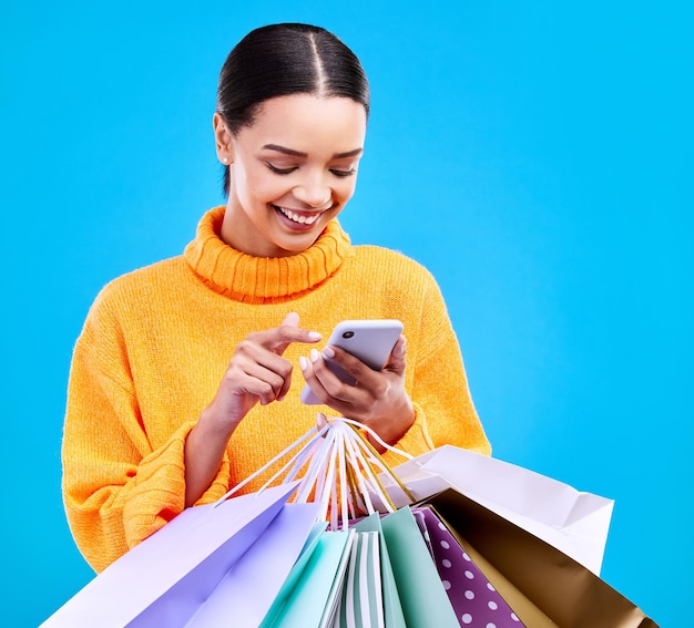 Teléfono de mujer feliz y bolsas de compras con una sonrisa para la compra, venta o descuento en un estudio azul
