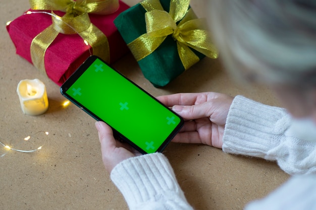 Un teléfono con una pantalla verde en manos de una mujer con un suéter blanco, una caja de regalo roja y verde y luces bokeh sobre la mesa. De cerca. Concepto de compras online para Navidad y año nuevo