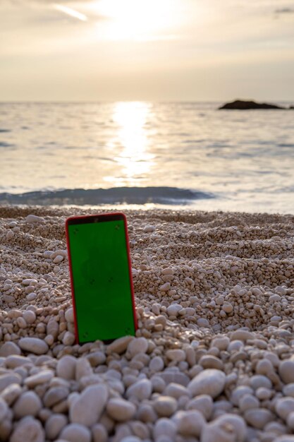 Teléfono con pantalla verde en la playa de mar rocoso en el espacio de copia al atardecer