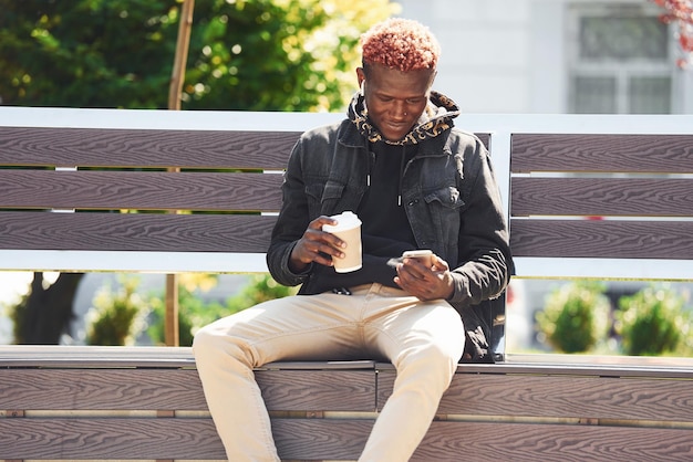 Con teléfono y taza de bebida Joven afroamericano con chaqueta negra al aire libre en la ciudad sentado en un banco