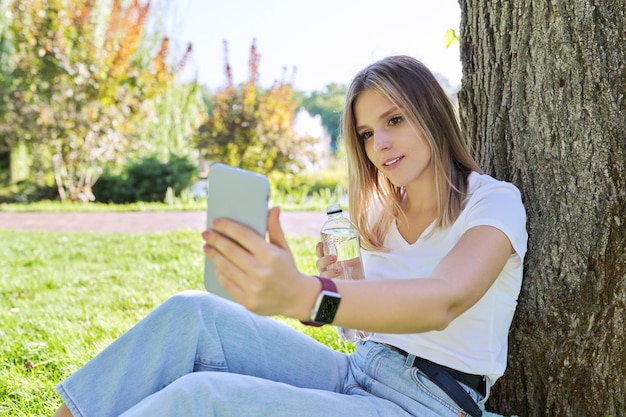 Telekommunikation Videoanruf Videokonferenz Kommunikation mit Freunden und Familie aus der Ferne Studentin Teenager Blick auf Smartphone-Bildschirm lachend reden auf Gras im Park sitzen