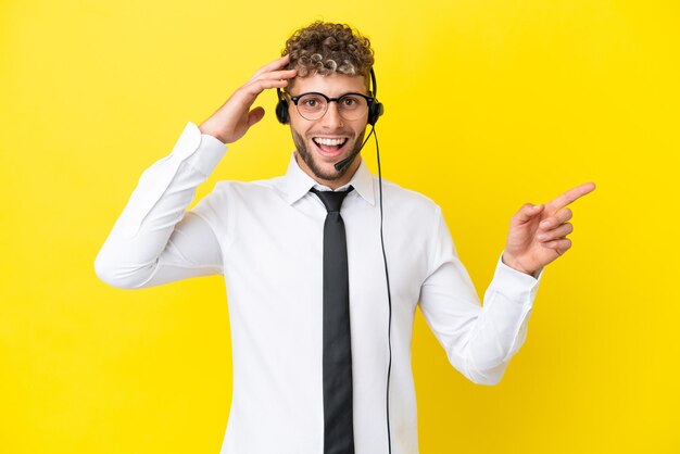 Telemarketer blonder Mann, der mit einem Headset arbeitet, das auf gelbem Hintergrund isoliert ist, überrascht und zeigt mit dem Finger zur Seite