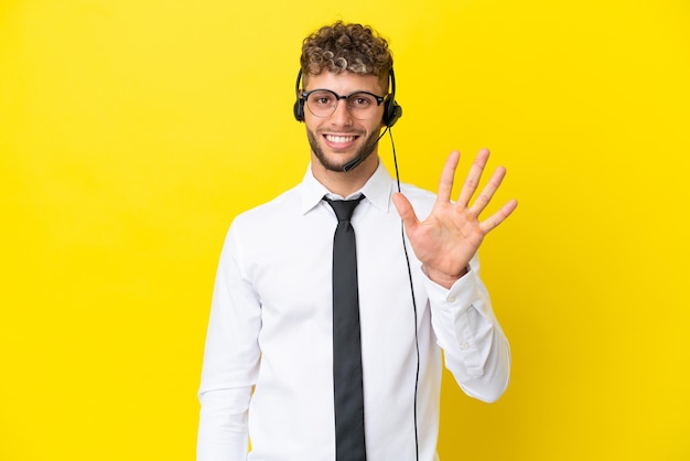 Telemarketer blonder Mann, der mit einem Headset arbeitet, das auf gelbem Hintergrund isoliert ist und fünf mit den Fingern zählt