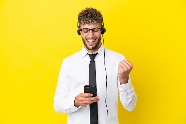 Telemarketer blonder Mann, der mit einem Headset arbeitet, isoliert auf gelbem Hintergrund mit Telefon in Siegesposition