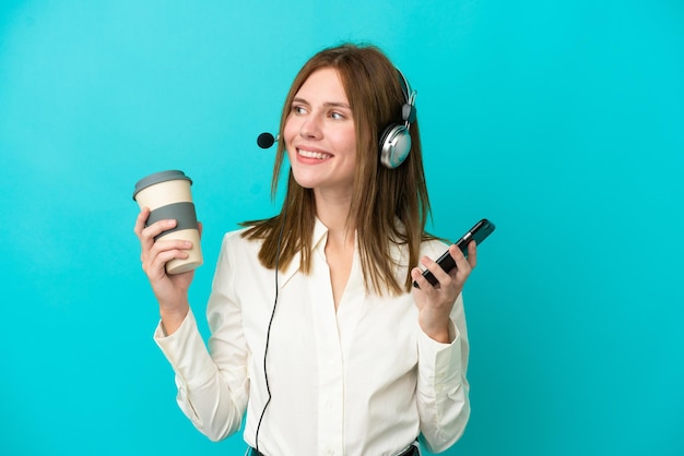 Telemarketer Engländerin, die mit einem Headset arbeitet, das auf blauem Hintergrund isoliert ist und Kaffee zum Mitnehmen und ein Handy hält