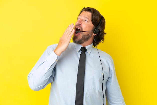 Telemarketer Holländer, der mit einem Headset arbeitet, das auf gelbem Hintergrund isoliert ist, gähnt und den weit geöffneten Mund mit der Hand bedeckt