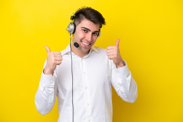 Telemarketer hombre caucásico que trabaja con un auricular aislado sobre fondo amarillo con pulgares arriba gesto y sonriendo