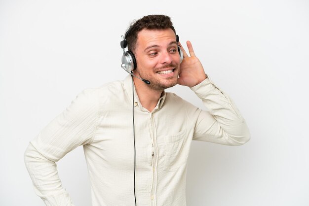 Telemarketer hombre caucásico que trabaja con un auricular aislado sobre fondo blanco escuchando algo poniendo la mano en la oreja