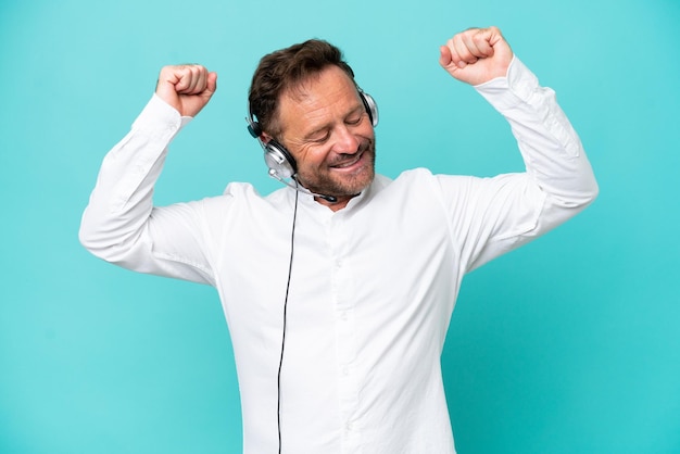 Telemarketer hombre caucásico trabajando con un auricular aislado sobre fondo azul celebrando una victoria