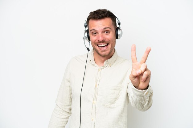 Telemarketer hombre caucásico trabajando con un auricular aislado sobre fondo blanco sonriendo y mostrando el signo de la victoria