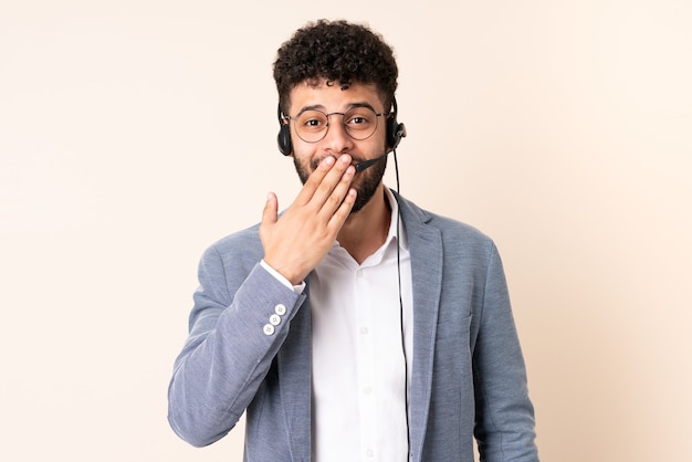 Telemarketer hombre marroquí que trabaja con un auricular aislado en la pared beige feliz y sonriente cubriendo la boca con la mano