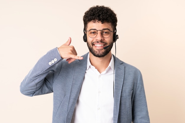 Telemarketer hombre marroquí que trabaja con un auricular aislado en la pared beige haciendo gesto de teléfono. Llámame señal