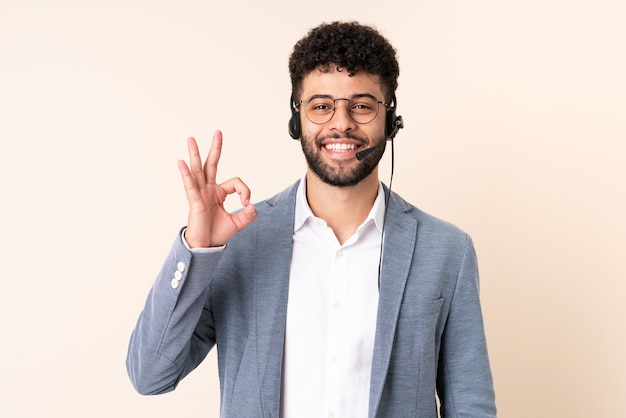 Telemarketer hombre marroquí que trabaja con un auricular aislado en la pared beige mostrando el signo de ok con los dedos