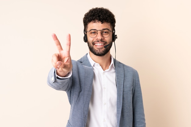 Telemarketer hombre marroquí que trabaja con un auricular aislado en la pared beige sonriendo y mostrando el signo de la victoria