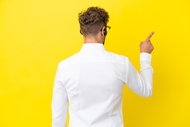 Telemarketer hombre rubio que trabaja con un auricular aislado sobre fondo amarillo apuntando hacia atrás con el dedo índice