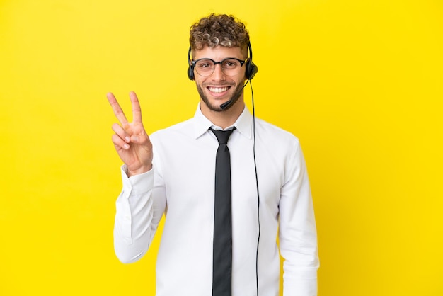 Telemarketer hombre rubio trabajando con un auricular aislado sobre fondo amarillo sonriendo y mostrando el signo de la victoria