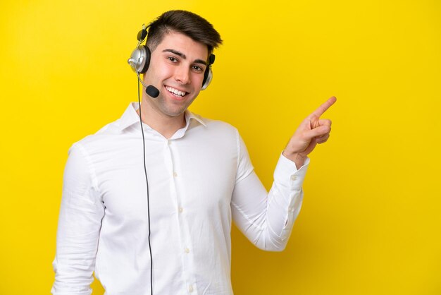 Telemarketer kaukasischer Mann, der mit einem Headset arbeitet, das auf gelbem Hintergrund isoliert ist und mit dem Finger zur Seite zeigt