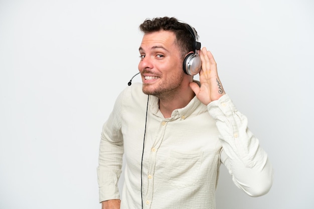 Telemarketer kaukasischer Mann, der mit einem Headset arbeitet, das auf weißem Hintergrund isoliert ist und etwas hört, indem er die Hand auf das Ohr legt