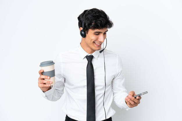 Telemarketer-Mann, der mit einem Headset arbeitet, das auf weißem Hintergrund isoliert ist und Kaffee zum Mitnehmen und ein Handy hält?
