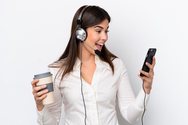 Telemarketer mujer caucásica que trabaja con un auricular aislado sobre fondo blanco con café para llevar y un móvil