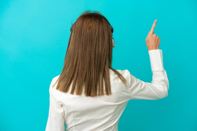 Telemarketer mujer inglesa que trabaja con un auricular aislado sobre fondo azul apuntando hacia atrás con el dedo índice