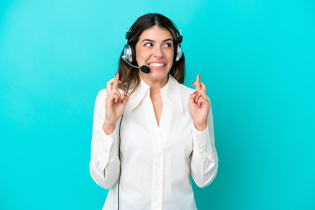 Telemarketer mujer italiana que trabaja con un auricular aislado sobre fondo azul con los dedos cruzados