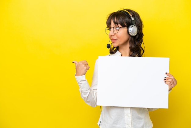 Telemarketer mujer latina que trabaja con un auricular aislado sobre fondo amarillo sosteniendo un cartel vacío y apuntando hacia el lado