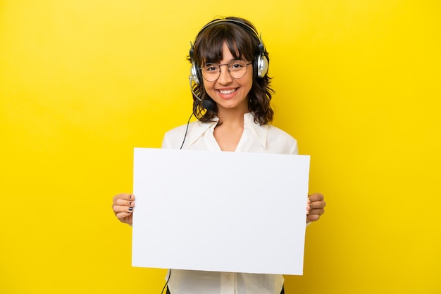 Telemarketer mujer latina trabajando con un auricular aislado sobre fondo amarillo sosteniendo un cartel vacío con expresión feliz