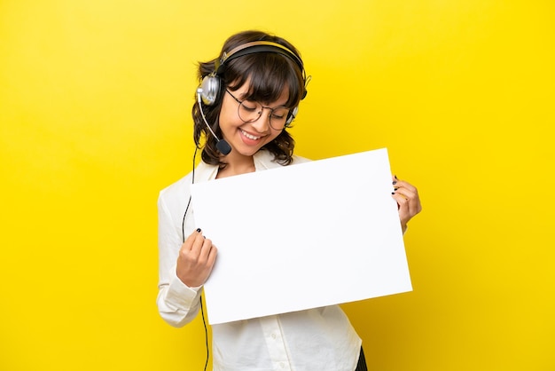 Telemarketer mujer latina trabajando con un auricular aislado sobre fondo amarillo sosteniendo un cartel vacío con expresión feliz