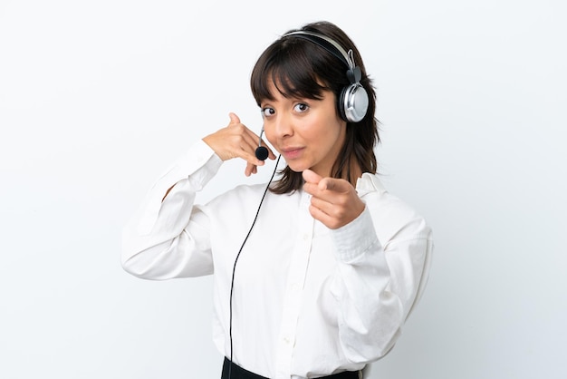 Telemarketer mujer de raza mixta que trabaja con un auricular aislado sobre fondo blanco haciendo gestos de teléfono y apuntando al frente