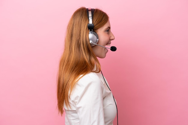 Telemarketer rothaarige Frau, die mit einem Headset arbeitet, das auf rosafarbenem Hintergrund isoliert ist und in Seitenlage lacht