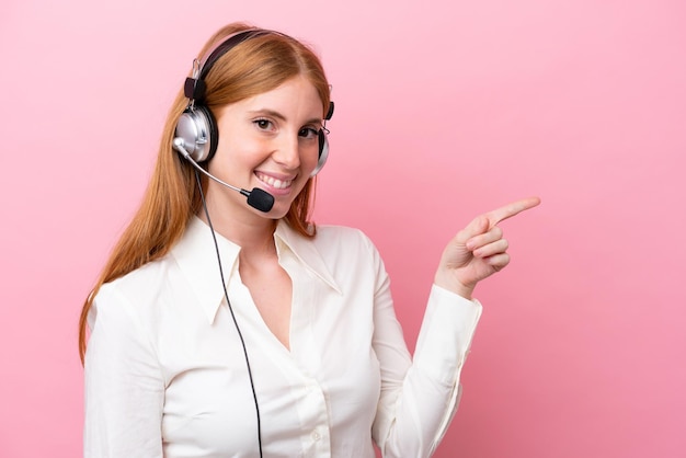 Foto telemarketer rothaarige frau, die mit einem headset arbeitet, das auf rosafarbenem hintergrund isoliert ist und mit dem finger zur seite zeigt