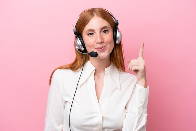 Telemarketer rothaarige Frau, die mit einem Headset arbeitet, das auf rosafarbenem Hintergrund isoliert ist und mit dem Zeigefinger zeigt, eine großartige Idee