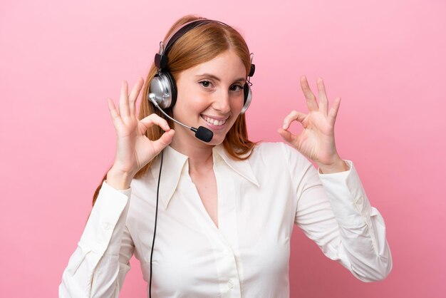 Telemarketer-Rothaarigefrau, die mit einem Headset arbeitet, das auf rosa Hintergrund isoliert ist und ein Ok-Zeichen mit den Fingern zeigt