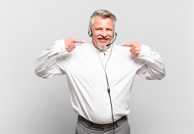 Telemarketer senior sonriendo con confianza apuntando a su propia sonrisa amplia, actitud positiva, relajada y satisfecha