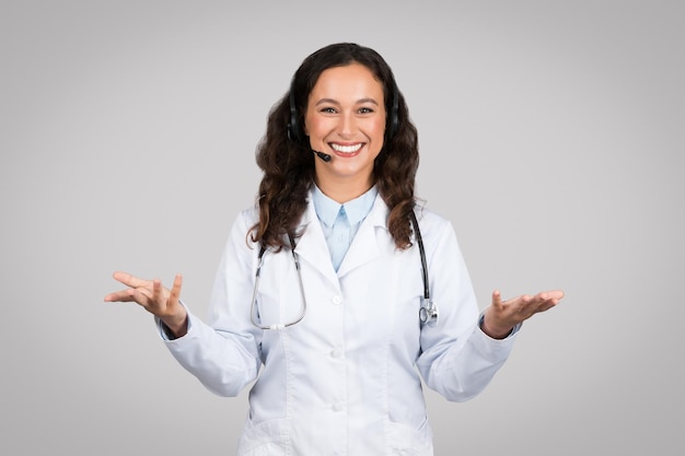 Telemedicina feliz mujer joven médico con auriculares y estetoscopio gesticulando y sonriendo a la cámara