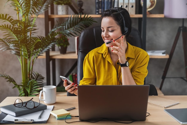 Teleoperatorin, die im Home Office mit Laptop und Headset arbeitet, während sie ihr Smartphone überprüft