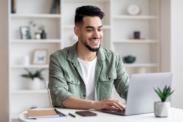 Teletrabalho feliz jovem árabe trabalhando remotamente com laptop no escritório em casa