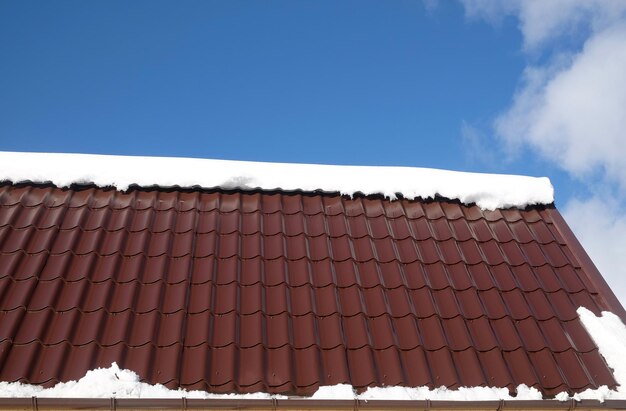 Telhado de telha de metal marrom com neve em um dia ensolarado de primavera sob um céu azul com nuvens brancas