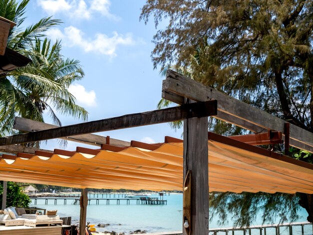 Telhado retrátil ao ar livre em construção de madeira e ferro no restaurante em frente à praia, perto da palmeira tropical e do fundo da vista marinha Toldo de lona dobrável à beira-mar