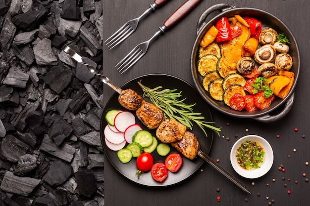 Teller mit Kebab und frischem Gemüse, Bratpfanne mit gegrilltem Gemüse auf einer schwarzen Holztischplatte mit Holzkohlehintergrund. Ansicht von oben.