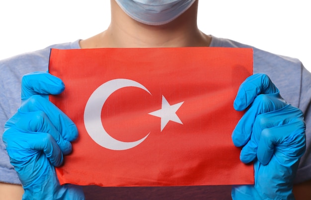 Tema pandémico del covid-19. Mujer con guantes protectores, mascarilla médica sostiene la bandera de Turquía aislada en blanco.