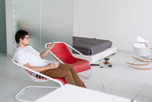 Tempo de lazer. homem de camisa branca sentado em uma cadeira assistindo tv, segurando uma xícara de chá e o telefone