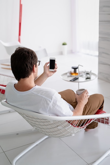 Tempo de lazer. Homem de camisa branca sentado em uma cadeira assistindo TV, segurando uma xícara de chá e o telefone