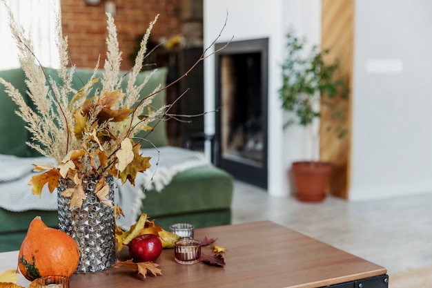 Foto tempo de outono decorado interior home. buquê de flores amarelas em um vaso de vidro na mesa de madeira elegante, abóbora, maçãs vermelhas, folhas, velas, cobertor. sofá verde, parede de tijolos e lareira no fundo